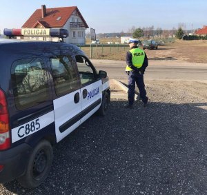 Policjant obserwuje ruch drogowy. Z lewej strony widoczny zaparkowany radiowóz.