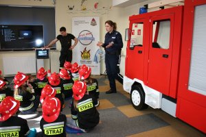 Policjantka  w siedzibie straży pożarnej omawia przepisy ruchu drogowego. Przed nią na podłodze siedzą dziec przebrane w stroje strażackie.