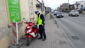 Miejsce zdarzenia drogowego. Policjant ruchu drogowego stoi na chodniku. obok niego z lewej strony motorower, który uczestniczył w kolizji. W oddali widać nadjeżdżające drogą krajową nr 5 pojazdy.