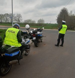 Dwóch umundurowanych policjantów podczas kontroli statycznej. Jeden z nich siedzi na  motocyklu, drugi stoi obok, mierząc prędkość nadjeżdżających pojazdów.