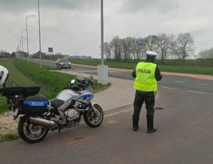 Umundurowany policjant  mierz  prędkość pojazdów ręcznym miernikiem prędkości, obok niego stoi motocykl.