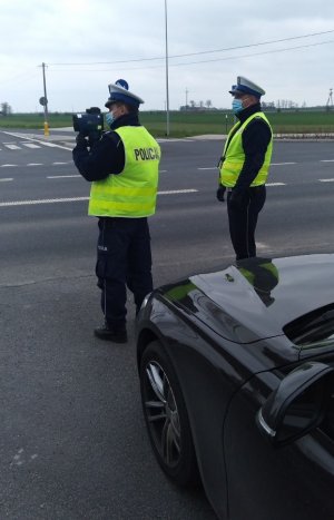 Dwóch policjantów  ruchu drogowego stojąc obok nieoznakowanego radiowozu mierzy prędkość.