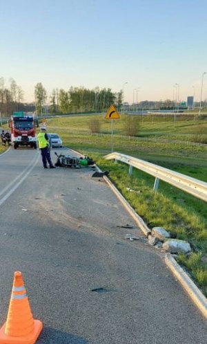 Na obrazku widoczny jest umundurowany policjant, uszkodzony motocykl oraz straż pożarna