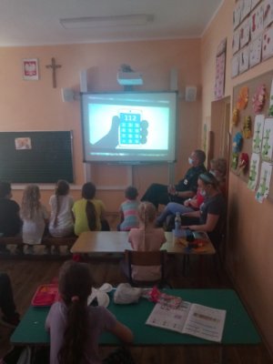 obrazek przedstawia dzieci w  klasie,  oraz informacje wyświetlane o numerach alarmowych na rzutniku