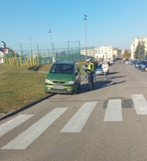 Policjanci wyjaśniają okoliczności potrącenia pieszej w Barcinie. Fotografia przedstawia miejsce zdarzenia. Na pierwszym planie widoczne przejście dla pieszych. W oddali policjant ruchu drogowego, który wykonuje czynność oględzin pojazdu marki Opel koloru zielonego, który brał udział w zdarzeniu.