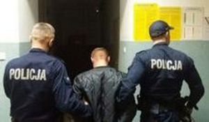 W holu komendy powiatowej stoi zatrzymany, obrócony tyłem. Po jego prawej i lewej stronie znajdują się policjanci, którzy trzymają go pod rękę.