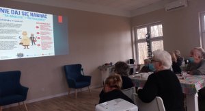 Wnętrze sali w Domu Pobytu Dziennego w Smerzynie. Z lewej strony stoły przy których siedzą seniorzy. z prawej strony na ścianie zamontowany rzutnik, na którym wyświetlany jest film poglądowy dot oszustw na policjanta i na wnuczka