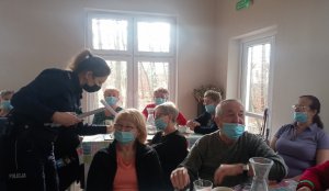 Sierż. szt. Katarzyna Leszczyńska - Krawczyk z lewej strony fotografii, która wręcza seniorom odblaski w kształcie pasków koloru zielonego. Z prawej strony widoczne dwa rzędu stołów, przy których siedzą seniorzy.