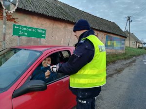 Policjant ruchu drogowego podczas kontroli. W czerwonym samochodzie siedzi kobieta. Z prawej strony policjant, który wręcza jej element odblaskowy i edukuje w zakresie obowiązujących przepisów.