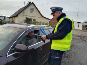 Policjant ruchu drogowego wręcza kierowcy ciemnego samochodu, który przewozi dzieci, kolorowe odblaski.