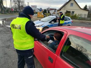 Policjant ruchu drogowego podczas czynności kontrolnych. Oddaje kierującej dokumenty. W oddali policyjny radiowóz i drugi funkcjonariusz.