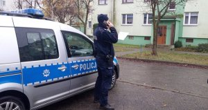 Policjant podczas sprawdzania kwarantanny. Z lewej strony stoi radiowóz. Przed policjantem blok wielorodzinny. Funkcjonariusz w ręku trzyma wykaz, a w drugiej ręce trzyma telefon.