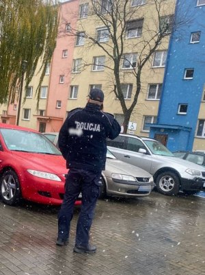 Kontrola kwarantanny. Policjant stoi przed blokiem. Przed nim zaparkowane samochody. Pada śnieg.