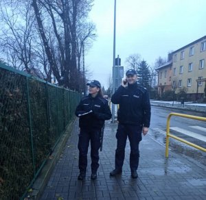 Policjanci podczas kontroli kwarantanny. Znajdują się na chodniku przy ulicy. Policjant rozmawia przez telefon, a policjantka obserwuje przez pobliski płot zachowanie osoby objętej kwarantanną.