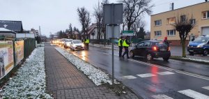 Widok ulicy Żnińskiej w Gąsawie. Pas drogowy, na którym z prawej i lewej strony widoczne jadące pojazdy. Na środku jezdni na obydwóch pasach ruchu stoją policjanci ubrani w żółte kamizelki. przy użyciu alkomatu sprawdzają trzeźwość kierowców.