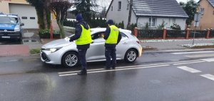 Na środku drogi stoją dwaj policjanci ubrani w kamizelki odblaskowe. Przed nimi na prawym pasie ruchu drogowego stoi biały samochód osobowy. Z lewej strony w oddali radiowóz.