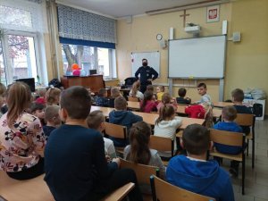 Sierżant sztabowy Arkadiusz Kaczmarek podczas prelekcji. Policjant stoi na środku klasy przy tablicy. Przed nim w ławkach siedzą dzieci.