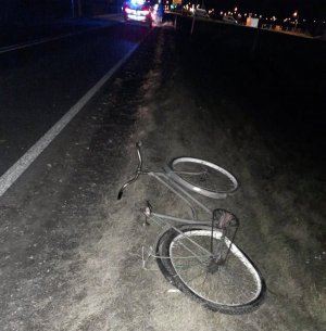 Panujący mrok. W sztucznym oświetleniu widoczna droga. Na poboczu na trawie po prawej stronie drogi leży uszkodzony rower, na którym nie ma odblasków ani oświetlenia. W oddali widać zaparkowany radiowóz policyjny.