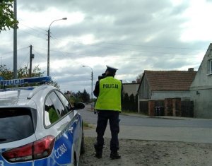 Policjant obserwuje ruch pojazdów. Policjant stoi tyłem. Z jego lewej strony znajduje się radiowóz. Z prawej strony widoczny budynek domu jednorodzinnego.