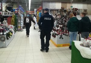 policjant  przemieszcza się po sklepie , pomiędzy regałami, znajdują się również osoby robiące zakupy