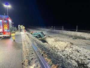 Miejsce zdarzenia drogowego.  Jest ciemno, miejsce oświetla halogen straży pożarnej. Z lewej strony widoczna trasa S5, a na niej stoi pojazd służbowy Straży pożarnej. przy nim z prawej strony stoją strażacy. Po środku znajdują się bariery energochłonne. Z prawej strony bariery energochłonnej rów, w którym na boku leży pojazd marki Citroen.