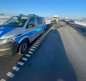 Widok na drogę S5. Po lewej stronie policyjny radiowóz, w którym siedzi policjant. W niewielkiej odległości za nim uszkodzony pojazd osobowy marki BMW koloru czarnego z uszkodzona szybą czołową. za nim pojazd ciężarowy marki Man. W oddali widoczne pojazd, które stoją ustawione w kolumnie. Tworzy się zator drogowy.