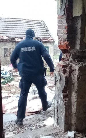 Widok z wnętrza pustostanu na podwórko. Funkcjonariusz wychodzi z tego budynku. Przed nim na podwórku widoczne rozrzucone sprzęty gospodarstwa domowego i zabawki, a także w centralnej części kolejny opuszczony budynek.