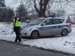 Okres zimowy. Z prawej strony na poboczu drogi stoi zaparkowany radiowóz przodem do jezdni. Obok stoi policjant ruchu drogowego, który w ręku trzyma tarczę do zatrzymywania pojazdów i daje nią sygnał do zatrzymania.