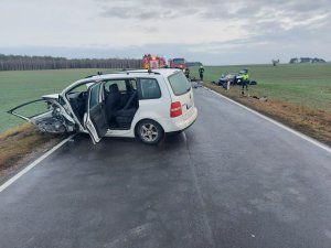 Na lewym pasie ruchu znajduje się samochód marki VW Touran koloru białego, na którym widoczne są uszkodzenia. W tle widoczne pojazdy straży pożarnej, które stoją na obydwu pasach ruchu. Z prawej strony pojazdów straży pożarnej znajdują się funkcjonariusze obsługujący zdarzenie. Z lewej i prawej strony fotografii pola uprawne.