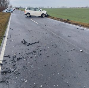 Widok drogi i miejsca w którym nastąpiło zderzenie. Na jezdni widoczne elementy karoserii, które oderwały się od pojazdów w chwili uderzenia. Wokół pola uprawne. W  oddali po prawej stronie pojazd marki VW touran koloru białego. Z lewej strony policyjny radiowóz oraz pojazdy, które czekają na możliwość przejazdu oraz osoby postronne.