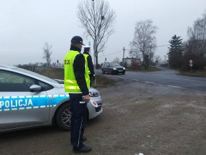 Policjanci obserwują ruch pojazdów. Z lewej strony stoi radiowóz, a przy nim z stoją funkcjonariusze. Jeden z nich w ręku trzyma tarcze do zatrzymywania pojazdów. Ubrani są w żółte kamizelki.