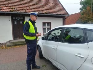 Policjant podczas kontroli drogowej. Za nim znajduje się budynek, a przed nim białe osobowe auto.