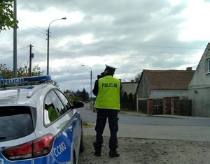 Z lewej strony zaparkowany policyjny radiowóz oznakowany. Obok niego po środku fotografii stoi policjant ruchu drogowego, który wykonuje pomiar prędkości. W oddali widoczne zabudowania oraz droga.