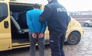 Policjanci podczas doprowadzenia zatrzymanego do sądu. Podejrzany na na rękach z tyłu kajdanki. Z jego prawej strony stoi policjant, drugi funkcjonariusz jest w środku pojazdu koloru żółtego, którym zatrzymany będzie przewożony.