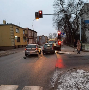 Miejsce kolizji, dwa pojazdy uczestniczące w zdarzeniu stoją na jezdni tuż przed sygnalizacją, przy  której stoi trzech pieszych. Za pojazdami osobowymi stoi karetka pogotowia.