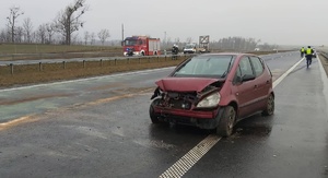 Miejsce zdarzenia. W centralnej części uszkodzony samochód. Za nim w oddali radiowóz i dwóch policjantów, którzy idą w jego kierunku. Z prawej strony za barierą energochłonną dwóch strażaków i ich pojazd służbowy.