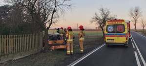 Z lewej strony ogrodzenie posesji i drzewo. przy nim stoi tyłem do autora zdjęcia pojazd marki Opel, którego tył jest uszkodzony w wyniku uderzenia w drzewo. Przy uszkodzonym pojeździe stoją strażacy i policjant. Z prawej strony na drodze zaparkowany ambulans, przed nim pojazd straży pożarnej.