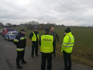 Zebranie komisji. W kółku stoją policjanci, biegły sądowy z dziedziny rekonstrukcji wypadków drogowych, funkcjonariusz Państwowej Straży Pożarnej, przedstawiciel Zarządu Dróg Powiatowych w Żninie z siedzibą w Podgórzynie. W tle pola uprawne i zaparkowane radiowozy.