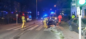 Widok miejsca zdarzenia od strony centrum miasta. Z prawej strony uszkodzone samochody, które uczestniczyły w zdarzeniu. W środkowej części rejon sygnalizacji świetlnej. Funkcjonariusz straży pożarnej zasypuje przy pomocy specjalnego preparatu wycieki płynów eksploatacyjnych na drodze.