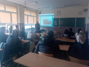 Policjantka w klasie lekcyjnej podczas omawiania tematyki dotyczącej doboru do służby w Policji. W ławkach siedzą uczniowie.