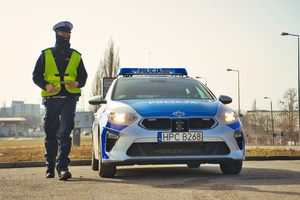 Policjant stoi obok radiowozu i obserwuje ruch pojazdów