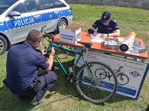 Dzielnicowi podczas znakowania rowerów. Z lewej strony radiowóz. Po środku fotografii stół przy którym siedzi policjantka i zapisuje znakowany numer. Przed nią specjalne oznaczenie na rower nanosi asp. Michał Starczewski.