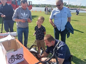 Dzielnicowy podczas znakowania roweru. Przy nim stoi chłopiec oraz trzech mężczyzn. W oddali Jezioro Małe Żnińskie oraz strażackie radiowozy.