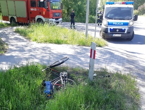 Fotografia przedstawiająca miejsce zdarzenia. na poboczu drogi po lewej stronie stoi wóz strażacki, z prawej strony policyjny radiowóz. w środkowej części w trawie na poboczu drogi leży rower.