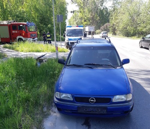 Samochód koloru niebieskiego, który brał udział w zdarzeniu drogowym. Za nim pojazd straży pożarnej i policyjny radiowóz. W rowie leży rower. Obok drogą jadą samochody.