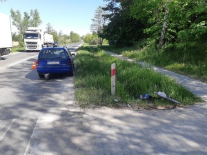 Widok miejsca zdarzenia. Z lewej strony droga jadą samochody. Po środku tyłem stoi niebieski samochód osobowy, na trawie leży uszkodzony rower. Na pierwszym planie widoczne przejście dla pieszych.