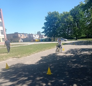 Dziewczynka na rowerze jedzie po specjalnie przygotowanym torze wyznaczonym przez żółte pachołki. Z lewej strony obserwuje ją dzielnicowy.