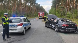 policjant  ruchu drogowego stoi na pasie  ruchu, gdzie  znajdują się uszkodzone  pojazdy i radiowóz policyjny