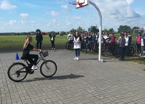 dziewczynka na  rowerze  pokonuje tor egzaminacyjny, na  płycie  boiska zgromadzona jest grupa uczniów z rowerami, nauczycielka oraz policjantka