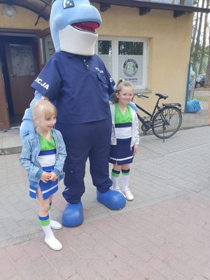 Przed budynkiem stadionu stoi Polfinek. Po jego obu stronach stoją dwie dziewczynki.